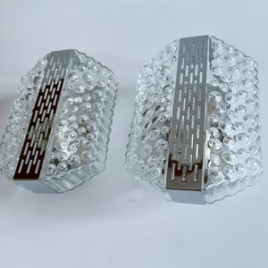 May include: Two wall sconces featuring clear, textured glass shades with a bubble-like pattern. Each fixture is accented by a central vertical chrome metal strip with a rectangular cutout design, providing a structured contrast to the organic glass.