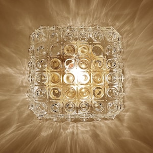 May include: A square flush mount ceiling light fixture featuring a clear glass shade with a grid of raised circular bubble patterns. The glass texture creates a warm, glowing light effect with distinct patterns projected onto the surrounding area.