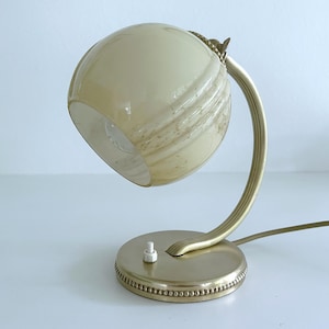 Może przedstawiać: Lampa stołowa z kulistym, kremowym szklanym kloszem z subtelnym brązowym marmurkowaniem. Lampa posiada zakrzywione, żebrowane metalowe ramię oraz okrągłą podstawę z koralikowym brzegiem w matowym złotym odcieniu i biały przełącznik dźwigienkowy.