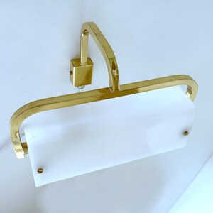 Può includere: Una lampada da parete per quadri con struttura in ottone lucido, braccio curvo e paralume rettangolare in acrilico bianco satinato. Il paralume è fissato alla struttura metallica tramite due piccole viti in ottone sulla parte anteriore.