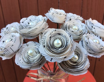May include: A bouquet of white paper flowers made from sheet music with pearl accents. The flowers are arranged in a clear glass vase.