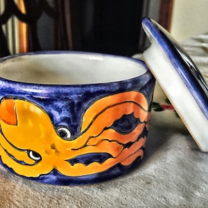 May include: A ceramic jar with a lid, featuring a hand-painted octopus design. The jar is cylindrical with a deep blue exterior and a bright orange octopus illustration. The lid is white with blue trim. The jar is likely used for storage.