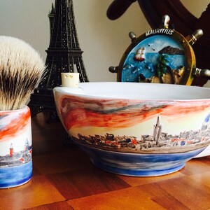 May include: A white ceramic shaving bowl with a colorful cityscape design. The bowl has a handle and is sitting on a wooden surface. A shaving brush with a brown and white bristle head is in a matching ceramic holder.