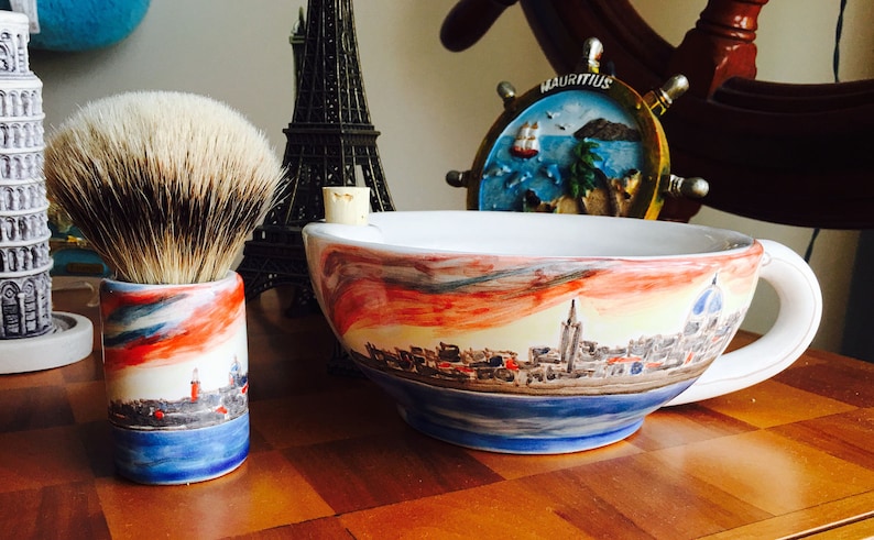 May include: A white ceramic shaving bowl with a colorful cityscape design. The bowl has a handle and is sitting on a wooden surface. A shaving brush with a brown and white bristle head is in a matching ceramic holder.