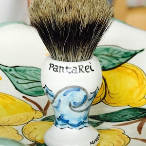 May include: A white ceramic shaving brush with a black and gray badger hair brush head. The base of the brush has a blue and white wave design and the words "Panta Rei" written in black. The brush is sitting on a ceramic tray with a lemon design.