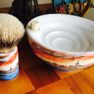 May include: A white ceramic shaving bowl with a cityscape design and a cork stopper. The bowl has a deep, round interior with a textured surface. A shaving brush with a white and brown bristle head is in a matching ceramic stand with a cityscape design.