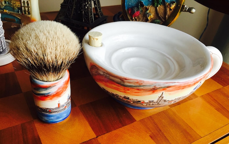 May include: A white ceramic shaving bowl with a cityscape design and a cork stopper. The bowl has a deep, round interior with a textured surface. A shaving brush with a white and brown bristle head is in a matching ceramic stand with a cityscape design.