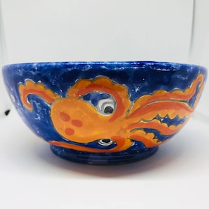 May include: A blue ceramic bowl with a hand-painted orange octopus design. The octopus has a large eye and tentacles.