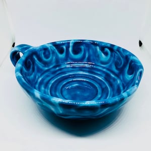 Op de afbeelding: Een handgemaakte blauwe keramische kom met een handvat. De kom heeft een wervelend patroon in blauwe en witte tinten. De kom is rond met een licht uitlopende rand en een klein handvat. De kom wordt waarschijnlijk gebruikt voor het serveren van voedsel.