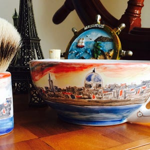 May include: A white ceramic shaving bowl with a painted cityscape design. The bowl has a handle and is decorated with a red, orange, and yellow sunset. The bowl is sitting on a wooden surface next to a shaving brush.