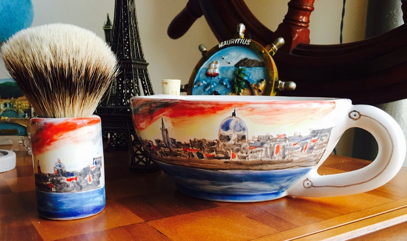 May include: A white ceramic shaving bowl with a painted cityscape design. The bowl has a handle and is decorated with a red, orange, and yellow sunset. The bowl is sitting on a wooden surface next to a shaving brush.