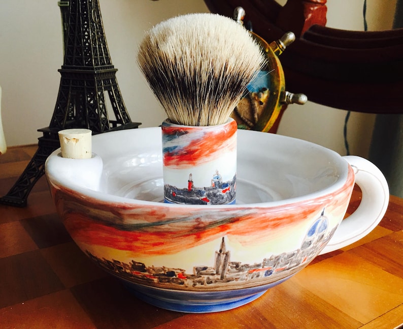 May include: A white ceramic shaving mug with a handle and a built-in soap dish. The mug has a painted cityscape design with red, orange, and blue colors. A brush with a white and gray bristle is in the soap dish.