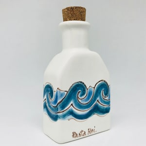May include: White ceramic bottle with a cork stopper and blue wave design. The bottle is decorated with a brown "Santa Rei" inscription.