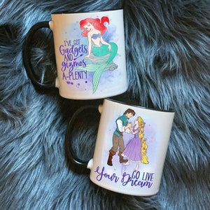 Pode incluir: Duas canecas de cerâmica branca com bordas pretas. A caneca à esquerda apresenta uma ilustração em aquarela de Ariel de A Pequena Sereia com o texto "Tenho gadgets e bugigangas de sobra". A caneca à direita apresenta uma ilustração em aquarela de Rapunzel e Flynn Rider de Enrolados com o texto "Viva seu sonho".