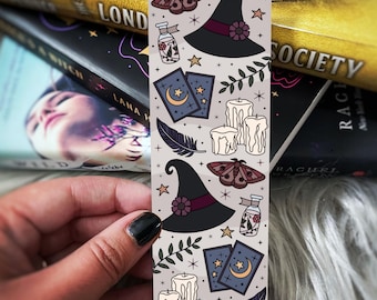 Puede incluir: Marcapáginas decorativo con ilustraciones de temática de brujas. El diseño incluye sombreros de bruja, polillas, velas, cartas del tarot y plumas, todo en una paleta de grises, negro y morado. El marcapáginas está sostenido en una mano, con libros en el fondo.