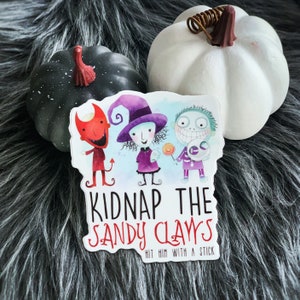 Pode incluir: Um adesivo com três personagens de desenhos animados do filme "O Estranho Mundo de Jack". Os personagens estão vestidos de bruxa, diabo e esqueleto. O adesivo diz "Kidnap the Sandy Claws" e "Hit him with a stick".