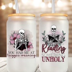 Può includere: Due bicchieri in vetro smerigliato con coperchi in bambù e cannucce in metallo. I bicchieri presentano uno scheletro che legge un libro con accenti floreali. Un bicchiere dice "You had me at trigger warning" e l'altro dice "Reading something unholy".