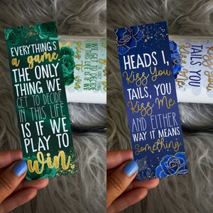 May include: Two bookmarks with gold lettering and floral designs. The green bookmark says "Everything's a game. The only thing we get to decide in this life is if we play to win." The blue bookmark says "Heads I, Kiss You. Tails, You Kiss Me. And either way it means something."