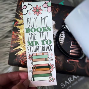 May include: A pink and green bookmark with the text "Buy me books and tell me to stfuatdlag" and a stack of books with flowers on the sides.