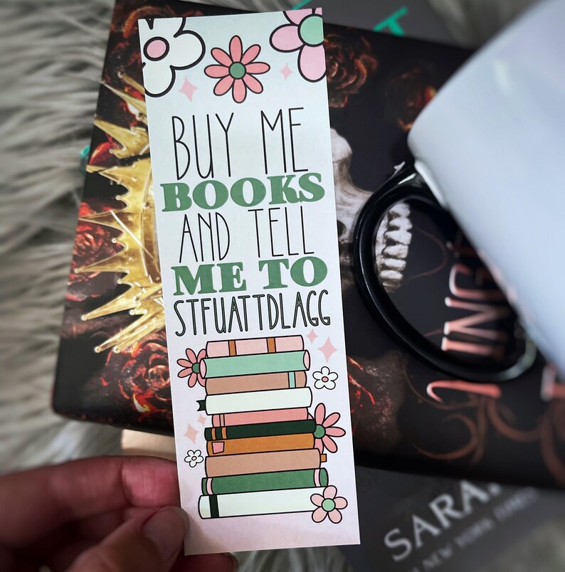 May include: A pink and green bookmark with the text "Buy me books and tell me to stfuatdlag" and a stack of books with flowers on the sides.