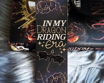May include: Black bookmark with gold lettering that reads "In My Dragon Riding Era". The bookmark features a black silhouette of a dragon with gold glitter accents and gold floral designs.