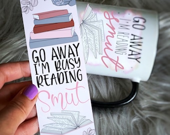 May include: A white bookmark with pink and black text that reads "Go Away I'm Busy Reading Smut". The bookmark features a stack of books, a rose, and an open book.