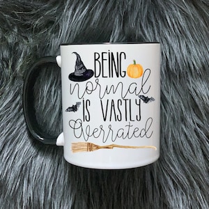 May include: White ceramic mug with a black rim and handle. The mug features a black witch's hat, a pumpkin, bats, and a broom with the text "Being normal is vastly overrated".