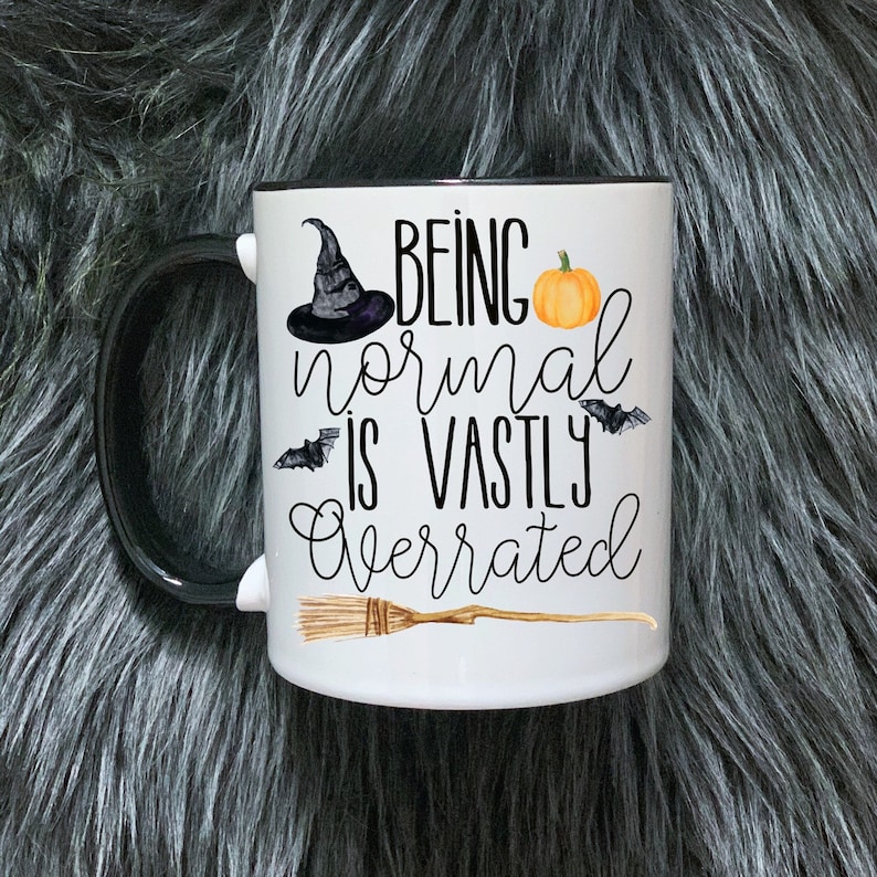 May include: White ceramic mug with a black rim and handle. The mug features a black witch's hat, a pumpkin, bats, and a broom with the text "Being normal is vastly overrated".