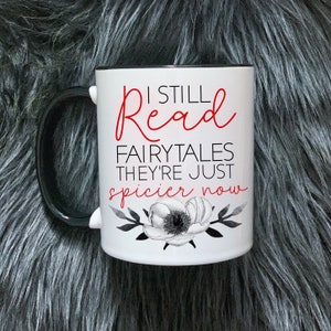 Może przedstawiać: Biały kubek ceramiczny z czarnym uchwytem. Kubek ma czerwony i czarny wzór z tekstem "I still read fairytales they're just spicier now".