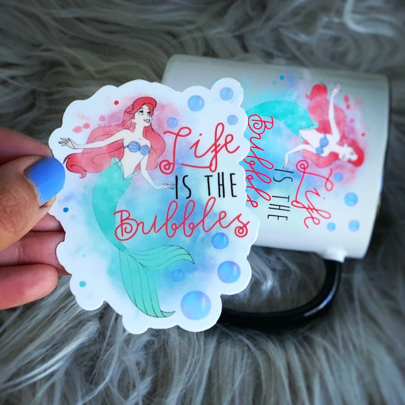 Puede incluir: Una taza de cer&aacute;mica blanca con asa negra que presenta una ilustraci&oacute;n de acuarela de Ariel de La Sirenita. La taza tiene el texto "Life is the Bubbles" impreso en ella.
