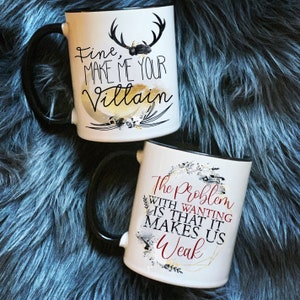Könnte beinhalten: Zwei weiße Keramikbecher mit schwarzen Henkeln und schwarzen Rändern. Der Becher links hat ein schwarzes und goldenes Design mit dem Text "Fine, Make Me Your Villain". Der Becher rechts hat ein schwarzes und goldenes Design mit dem Text "The Problem With Wanting Is That It Makes Us Weak".
