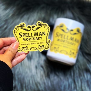 May include: Yellow sticker with black text that reads "Spellman Mortuary Funerals, Burials & Rites". The sticker is in a vintage style with a decorative border.