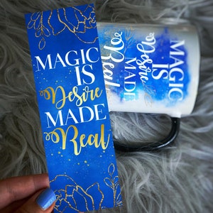 May include: A blue and gold bookmark with the text "Magic is Desire Made Real" on a blue background with gold glitter and stars. The bookmark is held in a hand with blue nail polish.