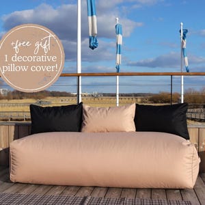 Könnte beinhalten: Ein hellrosa-farbener Sitzsack-Sofa mit schwarzen Kissenbezügen. Das Sofa steht auf einem Holzdeck mit Blick auf ein Gewässer und einen blauen Himmel. Der Text " +free gift 1 decorative pillow cover!" befindet sich auf einem Kreis in der oberen linken Ecke des Bildes.
