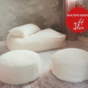 May include: A cozy off-white sherpa lounge set, including a large beanbag chair with a pillow and two round ottomans. The image features the text "Housewarming gift idea" in a red circle. The set is placed on a light wood floor.