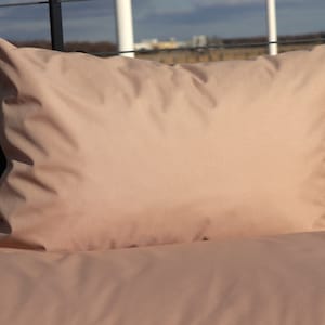 May include: A light brown pillow with a smooth, soft fabric surface. The pillow is lying on a bed with a matching light brown duvet cover.