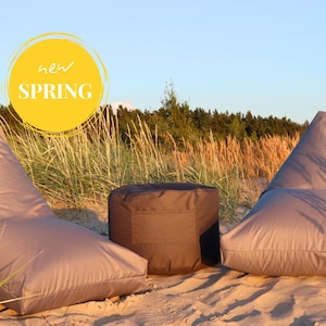 Può includere: Due poltrone a sacco grigie e un pouf rotondo grigio su una spiaggia sabbiosa con erba alta sullo sfondo. Un cerchio giallo con il testo "new SPRING" è nell'angolo in alto a sinistra dell'immagine.