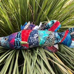 Może przedstawiać: Kolorowa ryba z patchworku wykonana z resztek tkaniny. Ryba jest niebieska, czerwona, zielona i biała. Ma duże oko i płetwę na grzbiecie. Ryba leży na łóżku z zielonej trawy.