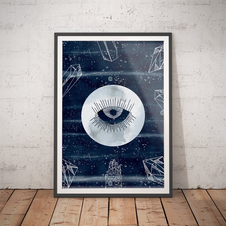 May include: A framed print with a watercolor background in shades of blue. The print features a white moon with a stylized eye in the center, surrounded by geometric shapes resembling crystals.