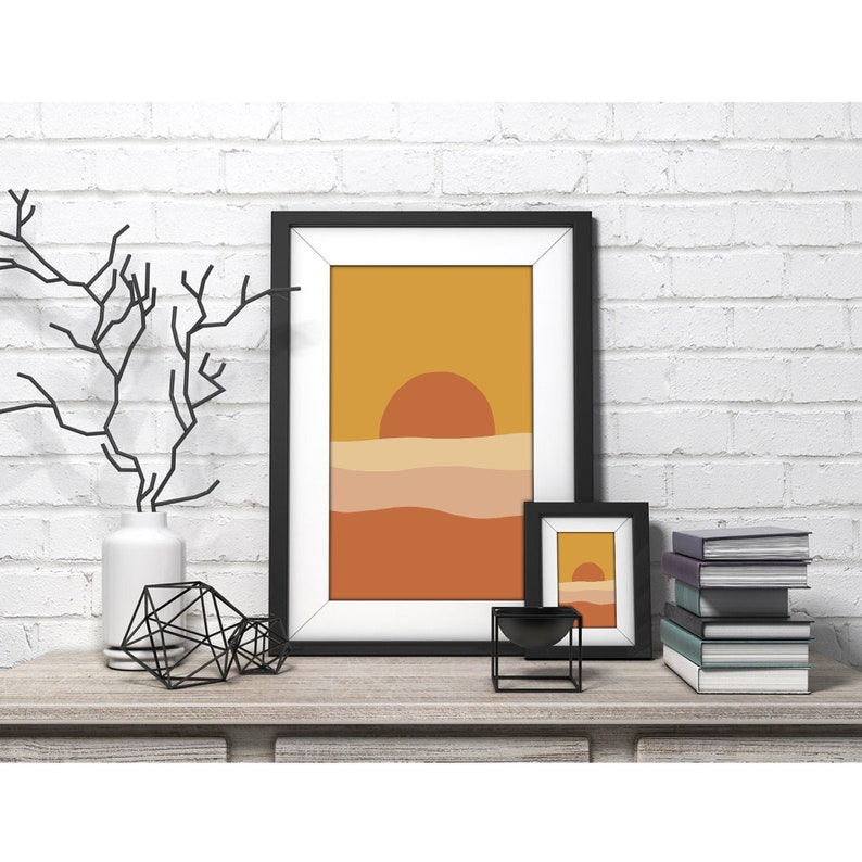 May include: Two framed abstract sunset prints with a minimalist design. The larger print features a yellow, orange, and brown color scheme. The smaller print is a smaller version of the larger print. The prints are displayed on a wooden shelf with a white vase, a black metal sculpture, and a stack of books.
