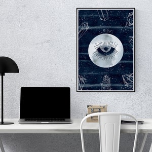 May include: A framed print with a dark blue background and a white moon with an eye in the center. The moon is surrounded by white geometric shapes and stars. The print is hanging on a white wall above a white desk with a black lamp and a laptop.