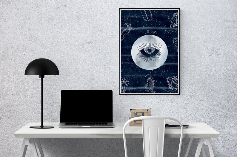 May include: A framed print with a dark blue background and a white moon with an eye in the center. The moon is surrounded by white geometric shapes and stars. The print is hanging on a white wall above a white desk with a black lamp and a laptop.