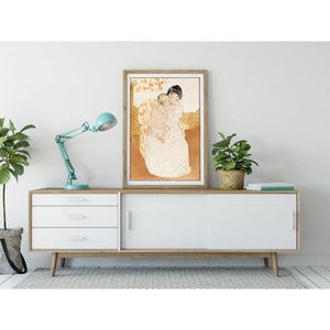 Op de afbeelding: Een ingelijste print van een schilderij dat een vrouw in een witte jurk toont die een kind vasthoudt. De vrouw en het kind zitten op een stoel met een bloemenpatroon. De achtergrond is licht oranje.