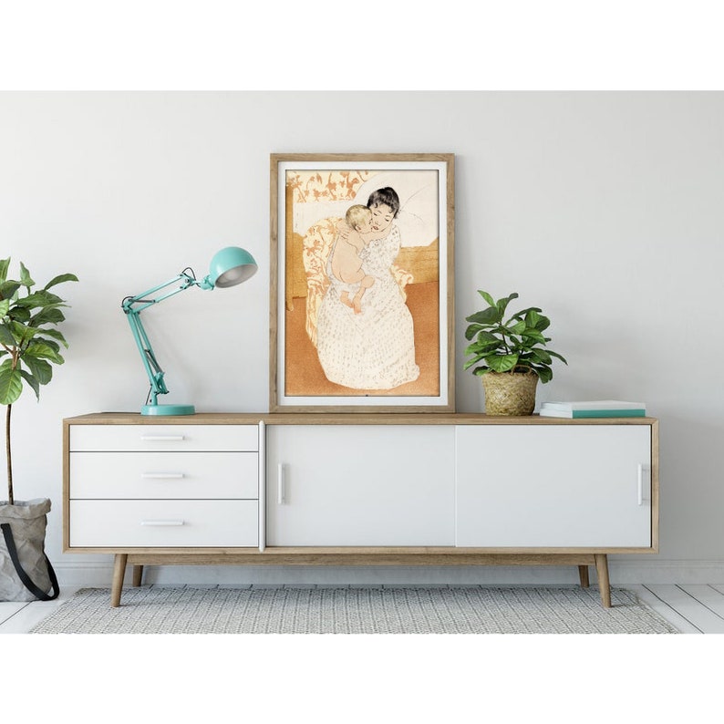Op de afbeelding: Een ingelijste print van een schilderij dat een vrouw in een witte jurk toont die een kind vasthoudt. De vrouw en het kind zitten op een stoel met een bloemenpatroon. De achtergrond is licht oranje.
