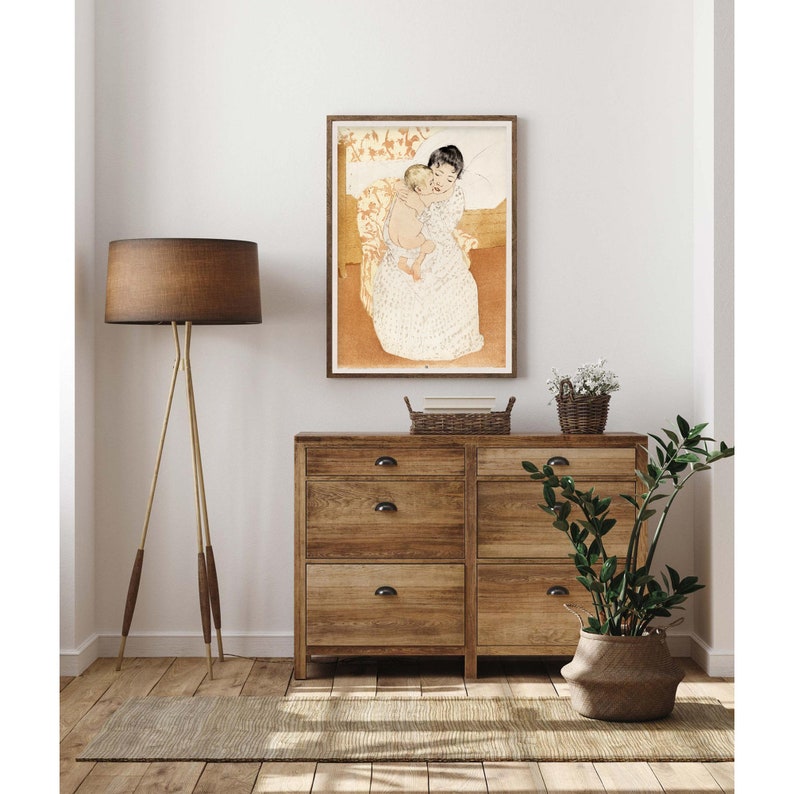 Op de afbeelding: Een vintage illustratie van een vrouw die een kind in haar armen houdt. De vrouw draagt een witte jurk met een bloemenpatroon en het kind draagt een witte jurk. De illustratie is in een lichtbruin frame en hangt aan een witte muur.