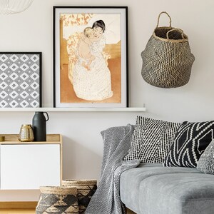 Op de afbeelding: Een ingelijste print van een schilderij dat een vrouw afbeeldt die een kind in haar armen houdt. De vrouw draagt een witte jurk met een bloemenpatroon en het kind is naakt. Het schilderij is geplaatst tegen een lichtbruine achtergrond.