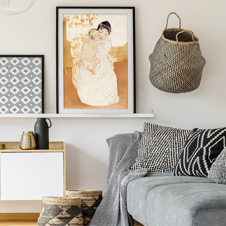 Op de afbeelding: Een ingelijste print van een schilderij dat een vrouw afbeeldt die een kind in haar armen houdt. De vrouw draagt een witte jurk met een bloemenpatroon en het kind is naakt. Het schilderij is geplaatst tegen een lichtbruine achtergrond.