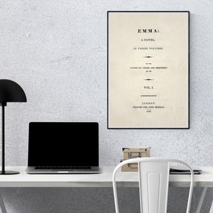 May include: A black and white framed print of the title page of the novel "Emma" by Jane Austen. The title page includes the text "Emma: A Novel in Three Volumes" and "Vol. I". The print is hanging on a white wall above a white desk with a black lamp, a laptop, and a stack of books.
