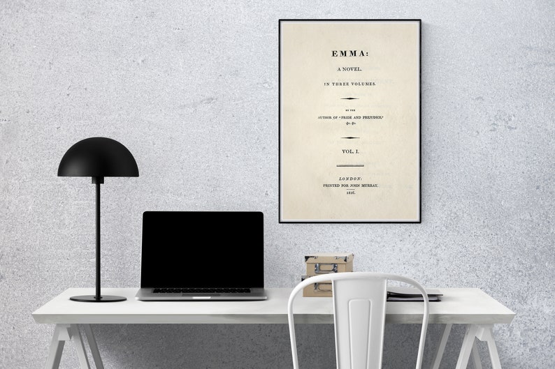 May include: A black and white framed print of the title page of the novel "Emma" by Jane Austen. The title page includes the text "Emma: A Novel in Three Volumes" and "Vol. I". The print is hanging on a white wall above a white desk with a black lamp, a laptop, and a stack of books.