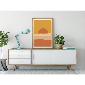 May include: A framed abstract print with a sunset scene in shades of orange, yellow, and brown. The print is hanging on a white wall above a white cabinet with wooden legs.